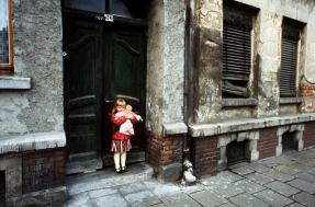 Ein kleines Mädchen steht am 12.3.1989 mit einer Puppe im Arm vor der Haustür eines maroden Wohnhauses in Leipzig © picture alliance / ZB | Waltraud Grubitzsch